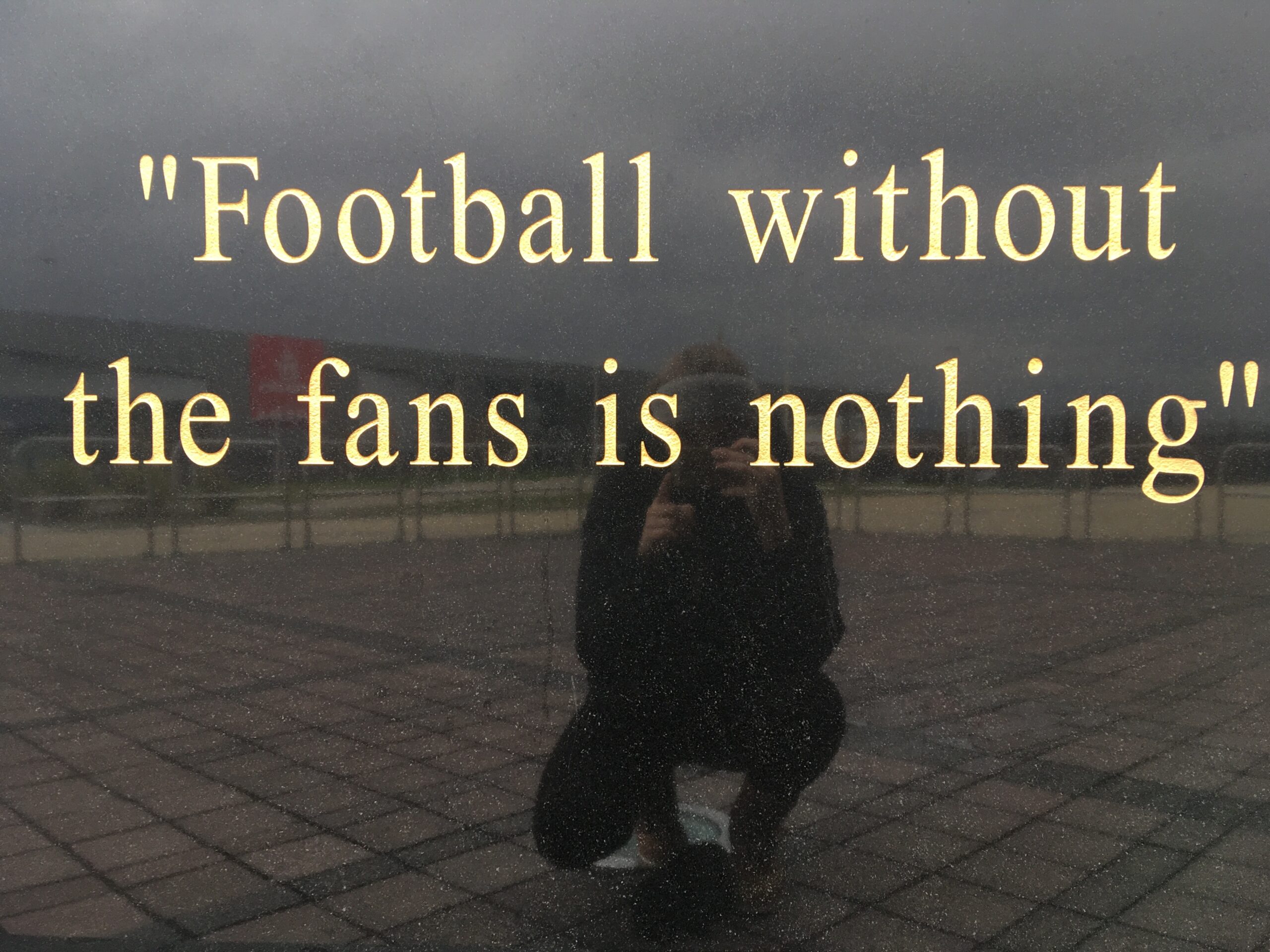 Zitat in goldener Schrift auf dunklem Hintergrund: „Football without the fans is nothing“. Im Spiegelbild darunter ist eine Person mit Kamera erkennbar, die das Foto auf einem Platz aufnimmt.