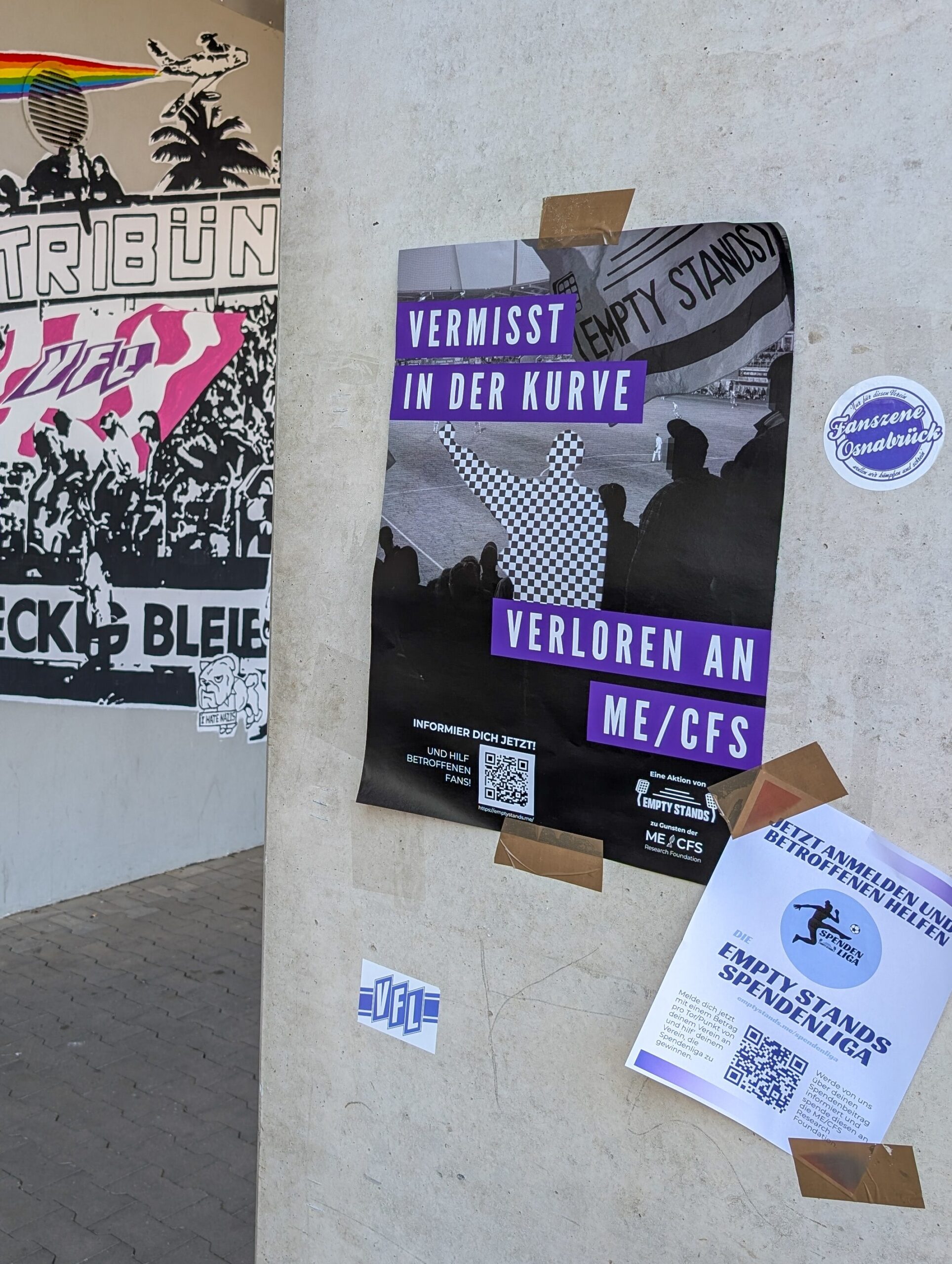 Straßenkunst und Plakate an einer Betonwand. Im Zentrum ein lila-schwarzes Plakat mit der Aufschrift 'VERMISST IN DER KURVE - VERLOREN AN ME/CFS', das Silhouetten von Fußballfans und einen QR-Code zeigt. Daneben verschiedene andere Aufkleber und Flyer. Unten rechts ein weißer Flyer mit blauem Text über Spendenliga.