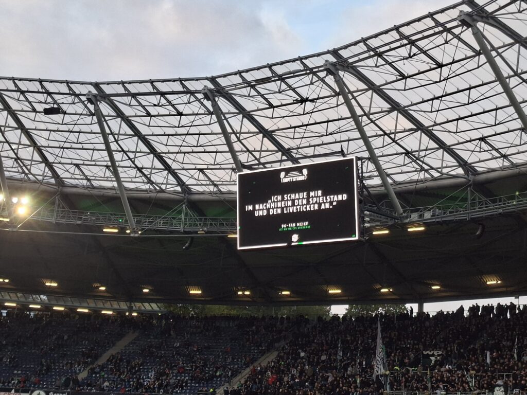 Bild der Leinwand der Heinz von Heiden Arena in Hannover. Auf der Leinwand ist oben das Empty Stands Logo abgebildet, darunter ein weißer Text auf schwarzem Hintergrund: "Ich schaue mir im Nachhinein den Spielstand und den Liveticker an", darunter "96-Fan Meike". Unter der Leinwand sind Oberränge zu sehen, darüber ein grauer Himmel.