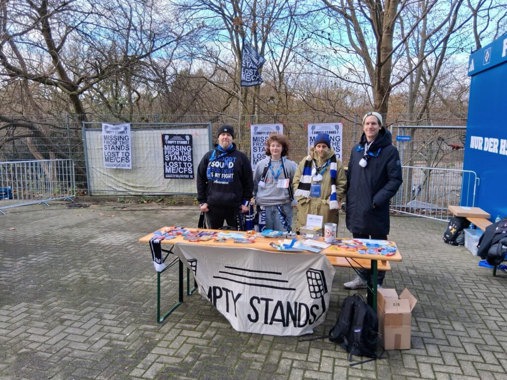 Vier Personen in Winterklamotten und Empty Stands sowie HSV-Schals stehen hinter einem Biertisch mit verschiedenen Infomaterialien, Flyern und Stickern. Davor hängt ein Banner mit dem schwarzen Logo von Empty Stands auf weißem Hintergrund. Im Hintergrund sind weitere schwarze und weiße Banner zu erkennen, auf denen steht "Missing from the Stands - Lost to ME/CFS", die an einem Bauzaun hängen. Dahinter sind blätterlose Bäume zu erkennen, am rechten Bildschirm ist ist eine blaue Wand zu erkennen, auf der in weißer Schrift "NUR DER HSV" steht.