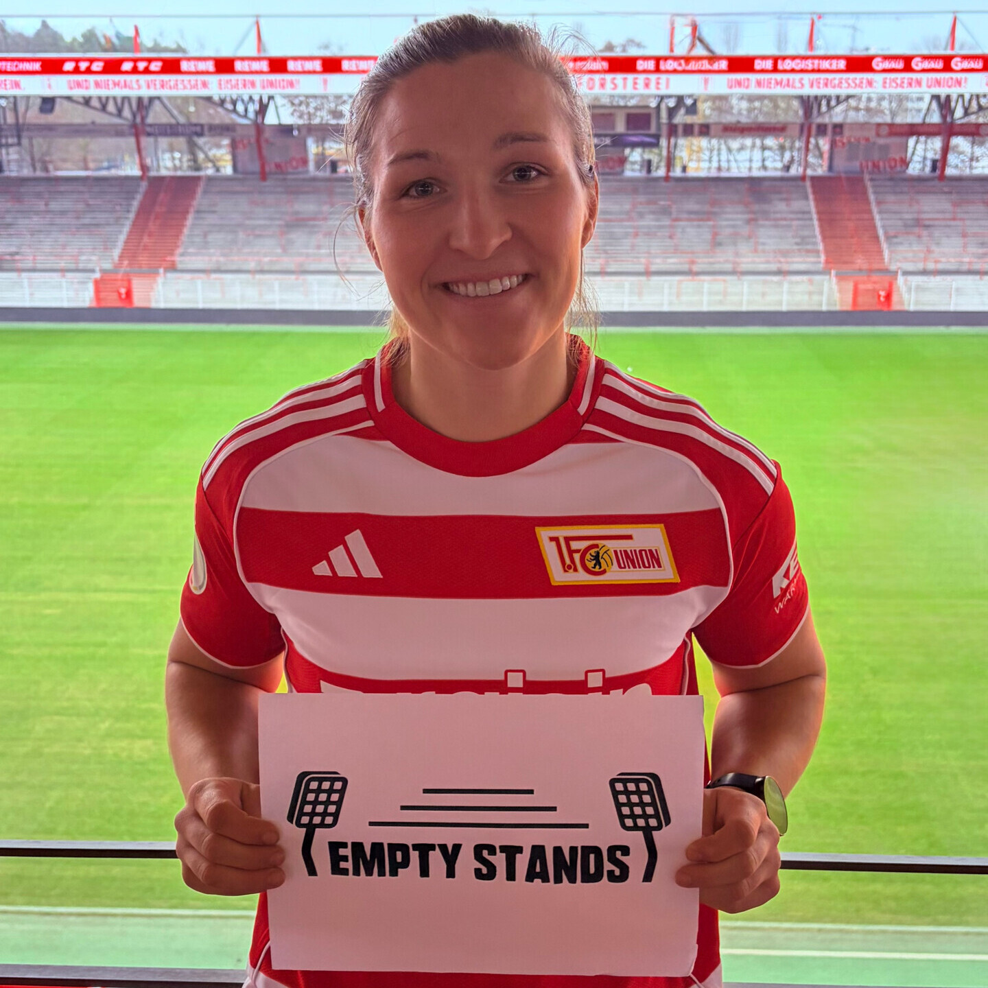 Eine Frau trägt ein rotes und weißes Trikot des 1. FC Union Berlin und hält ein Schild mit dem Schriftzug "EMPTY STANDS", flankiert von minimalistischen Flutlichtmasten und horizontalen Linien, die leere Ränge symbolisieren. Im Hintergrund ist ein leerer Fußballstadionplatz mit grünem Rasen und roten Tribünen zu sehen.