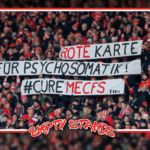 Bild einer Fankurve. Die Fans halten Banner hoch, auf denen steht: "Rote Karte für Psychosomatik! #CUREMECFS". "MECFS" und "Rote" sind rot eingefärbt, während der Rest schwarz eingefärbt ist.