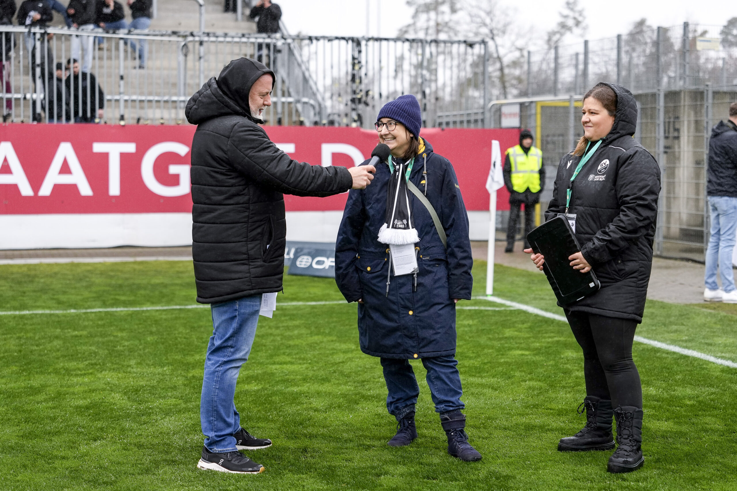 Drei Personen auf einem Fußballplatz. Ein Mann in schwarzer Pufferjacke und Jeans führt ein Interview mit einer Frau in blauem Mantel, dunkelblauer Mütze und einem schwarz-weißen Schal mit der Aufschrift. Eine weitere Frau in schwarzer Kapuzenjacke hält ein Tablet. Im Hintergrund ist eine rote Werbebande mit der Aufschrift "AATG D", eine Person in gelber Warnweste sowie Zuschauer*innen in den Tribünen zu sehen. Die Szene spielt auf einem grünen Rasen unter bewölktem Himmel.