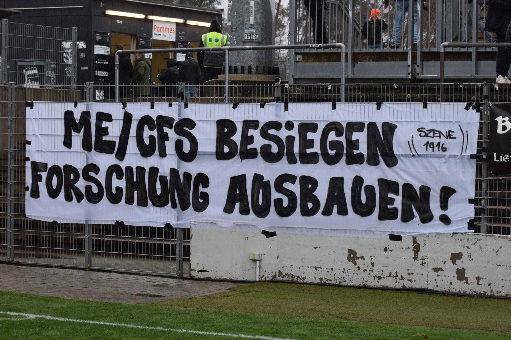 Ein weißes Spruchband mit schwarzer Schrift hängt an einem Metallzaun vor einem Fußballstadion. Der Text lautet: "ME/CFS BESIEGEN FORSCHUNG AUSBAUEN!" sowie "SCENE 1916" in kleinerer Schrift rechts oben. Das Spruchband ist ein Protest der Selbsthilfegruppe "Empty Stands" für Fußballfans mit ME/CFS, Long COVID & Post-Vac. Im Hintergrund sind Menschen an einem Zaun zu sehen, darunter ein Gebäude mit Schildern wie "Pommes" und "BECKS". Der Boden ist mit Rasen und einem gepflasterten Bereich versehen.