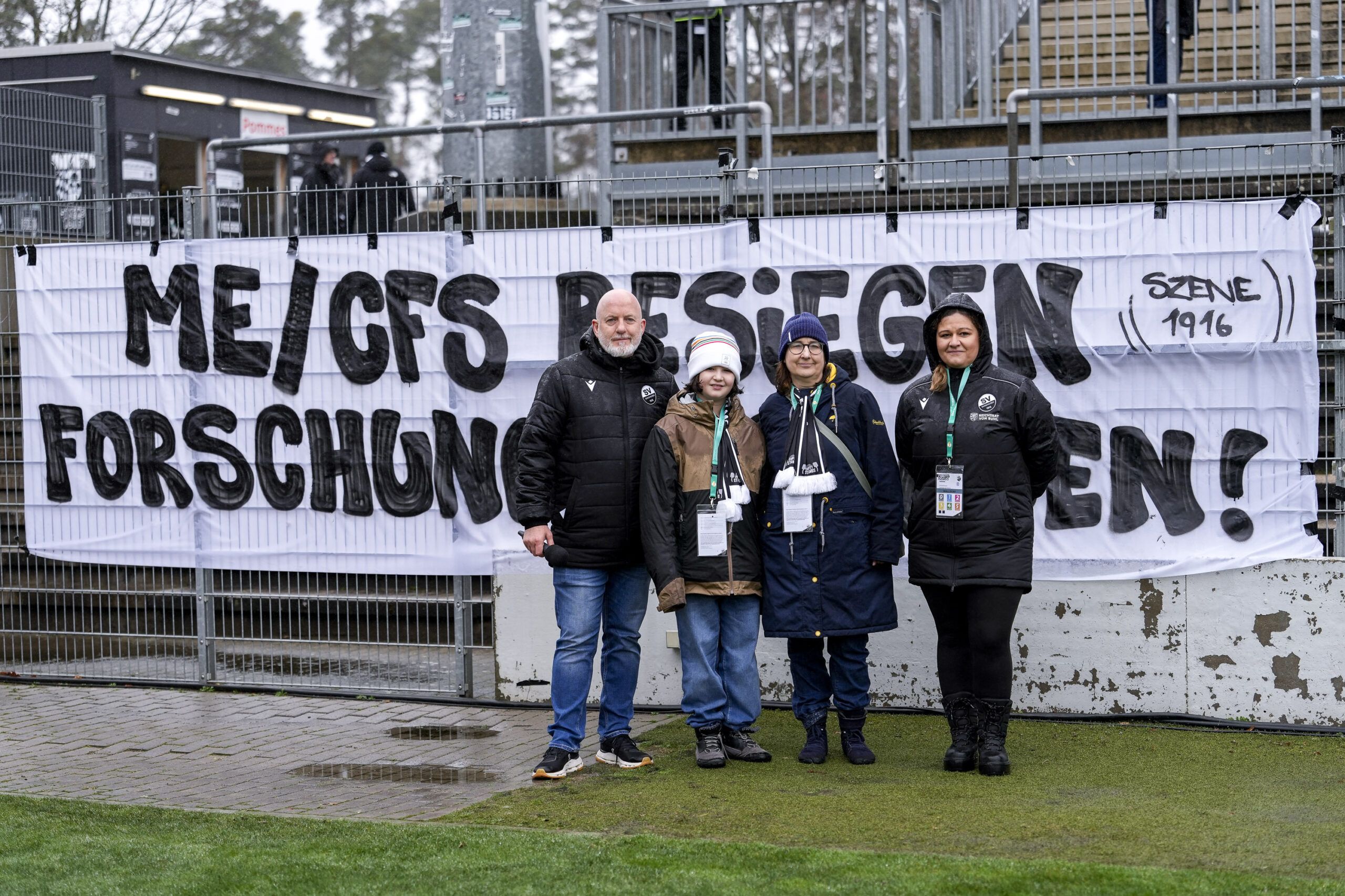 Vier Personen stehen vor einem weißen Banner mit der Aufschrift "ME/CFS BESEGEN FORSCHUNG DEN! (SZENE 1916)" in einem Stadionbereich mit Gras, Zäunen und Gebäuden im Hintergrund. Die Gruppe besteht aus Fußballfans mit ME/CFS, Long COVID & Post-Vac. Die Personen tragen Jacken und Mützen, der Hintergrund zeigt eine offene Tribüne mit leerem Stadionbereich.