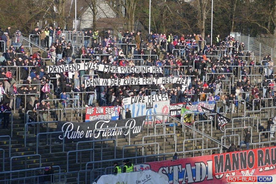 Bild einer Stehtribüne des Stadios am Zoo in Wuppertal. Die Ränge sind teilweise leer, in der Mitte gut gefüllt. Die Fans halten Banner hoch, auf denen steht: "Dringend muss mehr Forschung her, denn in der Kurve fehlt heut wer # CURE ME/CFS". Vorne sind weitere Banner in rot-blau zu erkennen.