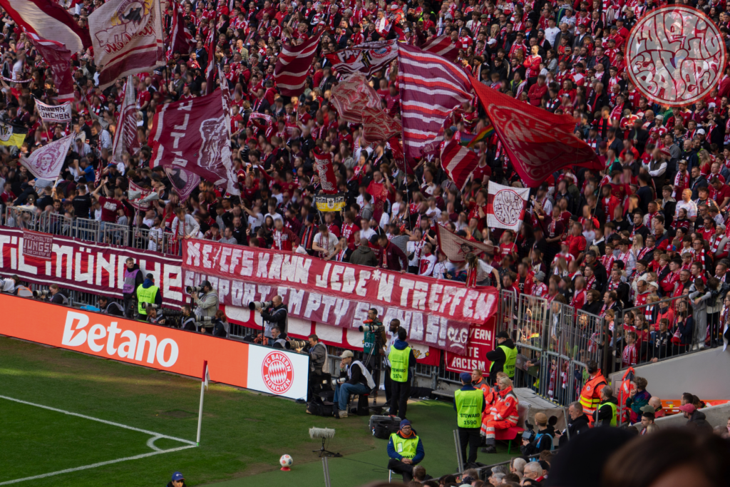 Generiert: Eine große Menschenmenge in einem Fußballstadion, die überwiegend in Rot und Weiß gekleidet ist. Zahlreiche Banner und Fahnen sind zu sehen, die eine intensive Fanatmosphäre erzeugen. Im Vordergrund ist der Rasenplatz zu erkennen.