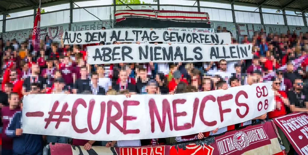 Generiert: Eine große Menschenmenge von Fußballfans in einem Stadion. Im Vordergrund halten die Fans ein großes Banner mit der Aufschrift „AUS DEM ALLTAG GEWORFEN, ABER NIEMALS ALLEIN“ und darunter ein weiteres Banner mit dem Hashtag „#CURE ME/CFS“. Die Fans demonstrieren damit ihre Solidarität und fordern Forschung für die Krankheit ME/CFS.