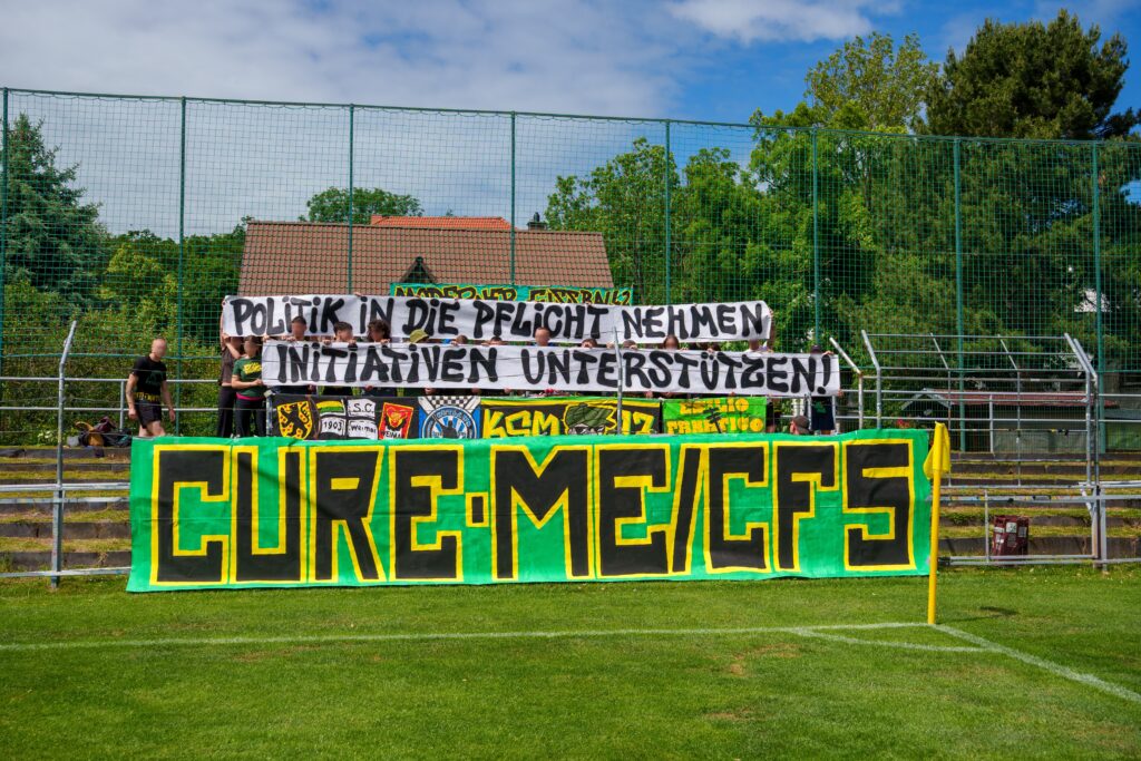 Generiert: Vor einem leeren Fußballtor auf einem Rasenplatz halten mehrere Personen große Banner. Das größte Banner trägt in großen gelben Buchstaben die Aufschrift „CURE ME/CFS“. Darüber sind kleinere Banner angebracht, die die Forderungen „POLITIK IN DIE PFLICHT NEHMEN“ und „INITIATIVEN UNTERSTÜTZEN“ tragen.